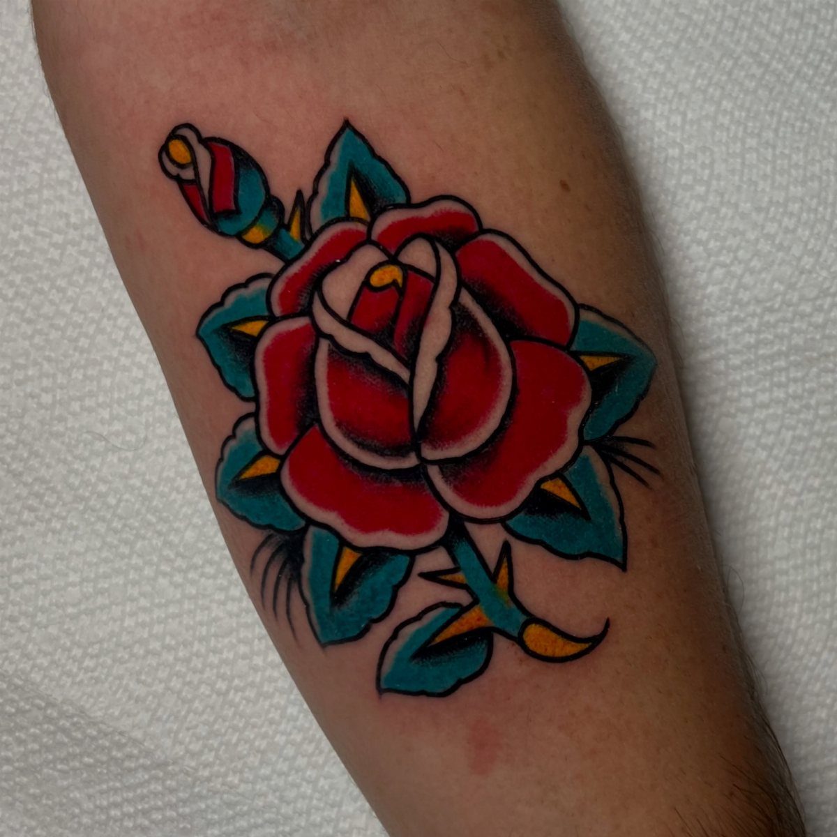 A red rose tattoo with teal leaves on a person's arm.