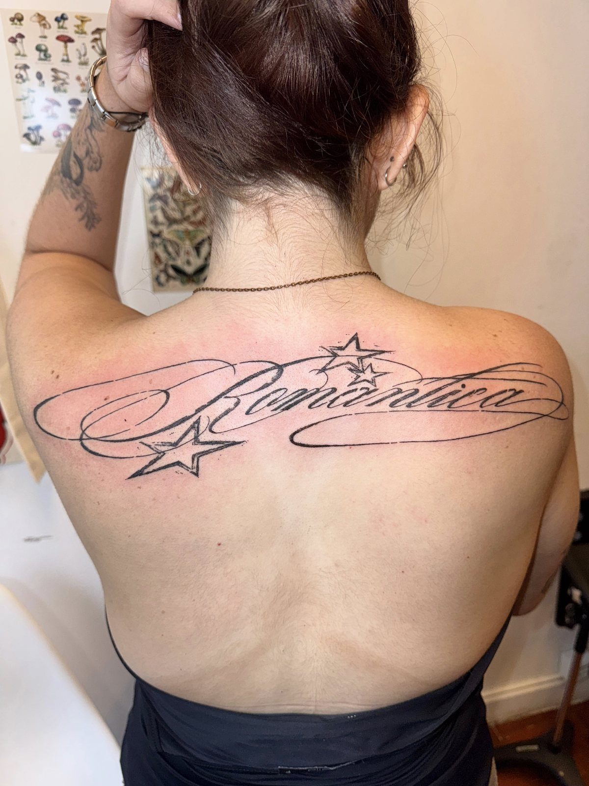7 Back Tattoo Writing Ideas for Lettering That Lasts 4 A woman's upper back displaying a cursive tattoo of the word Romantica with stars and swirls.