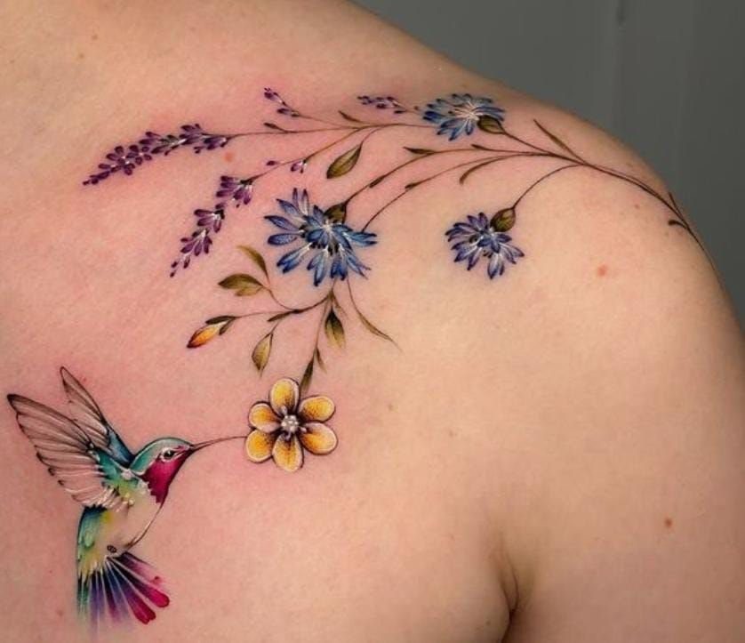 A colorful hummingbird tattoo hovering near a yellow flower and delicate blue floral branches on skin.