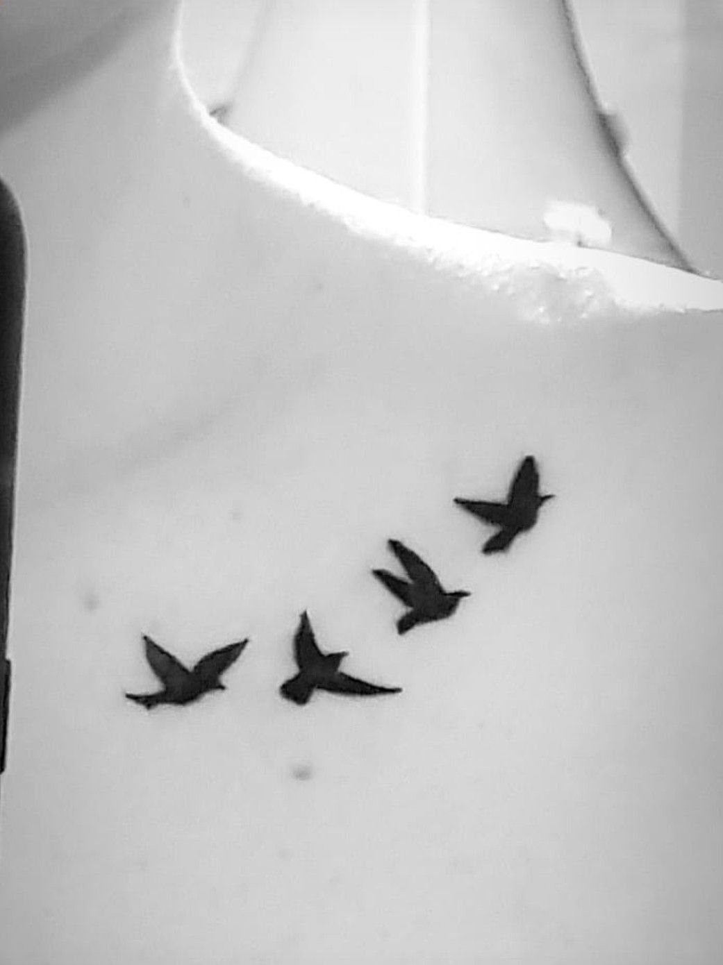 Four small black silhouette bird tattoos flying across a person's collarbone in black and white.