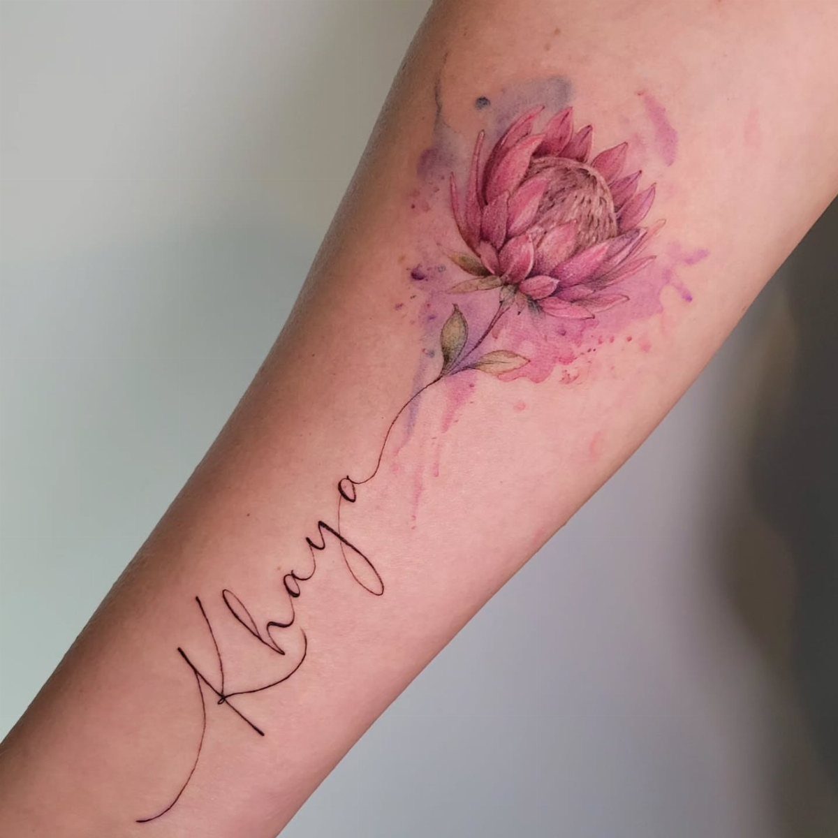 A pink watercolor protea flower tattoo with cursive script on a person's arm.