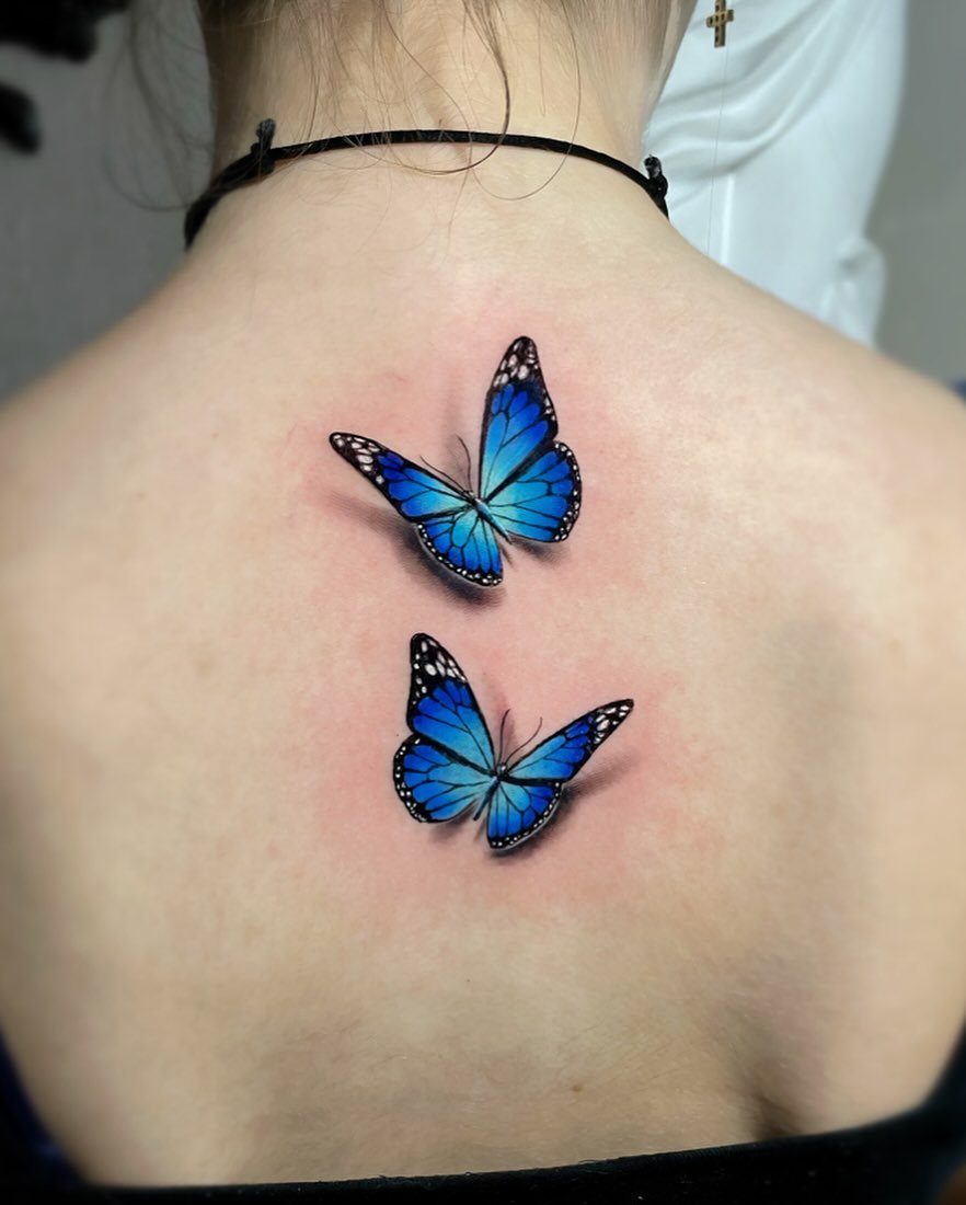 Two realistic blue butterfly tattoos with shadows on a person's upper back.