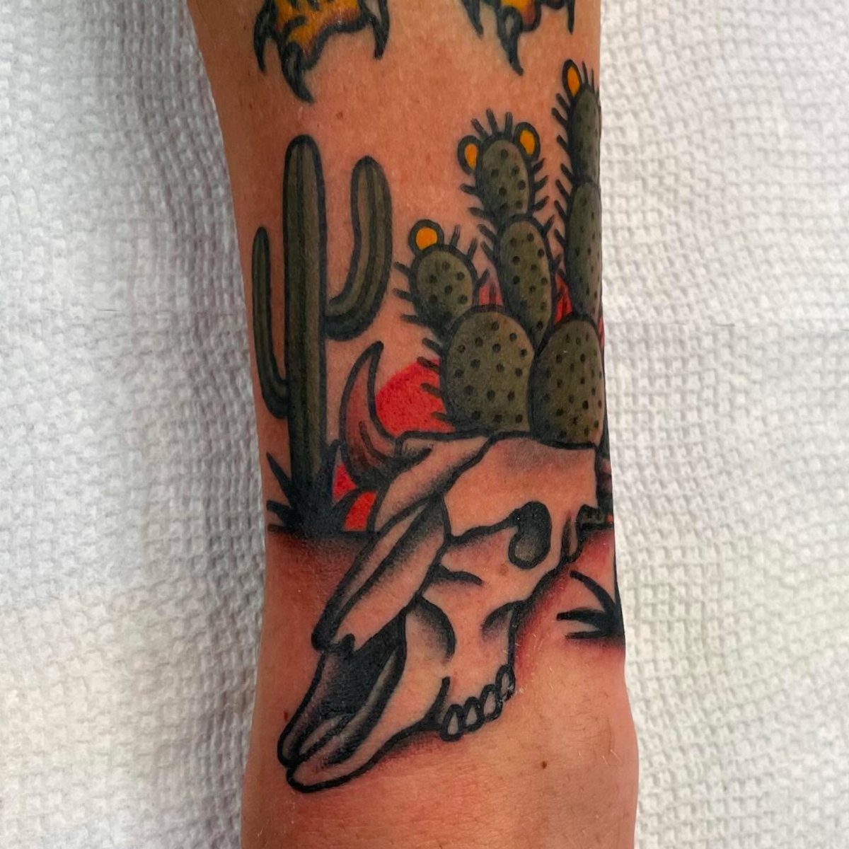 A tattoo of a cow skull with green cacti and a red sun on skin.