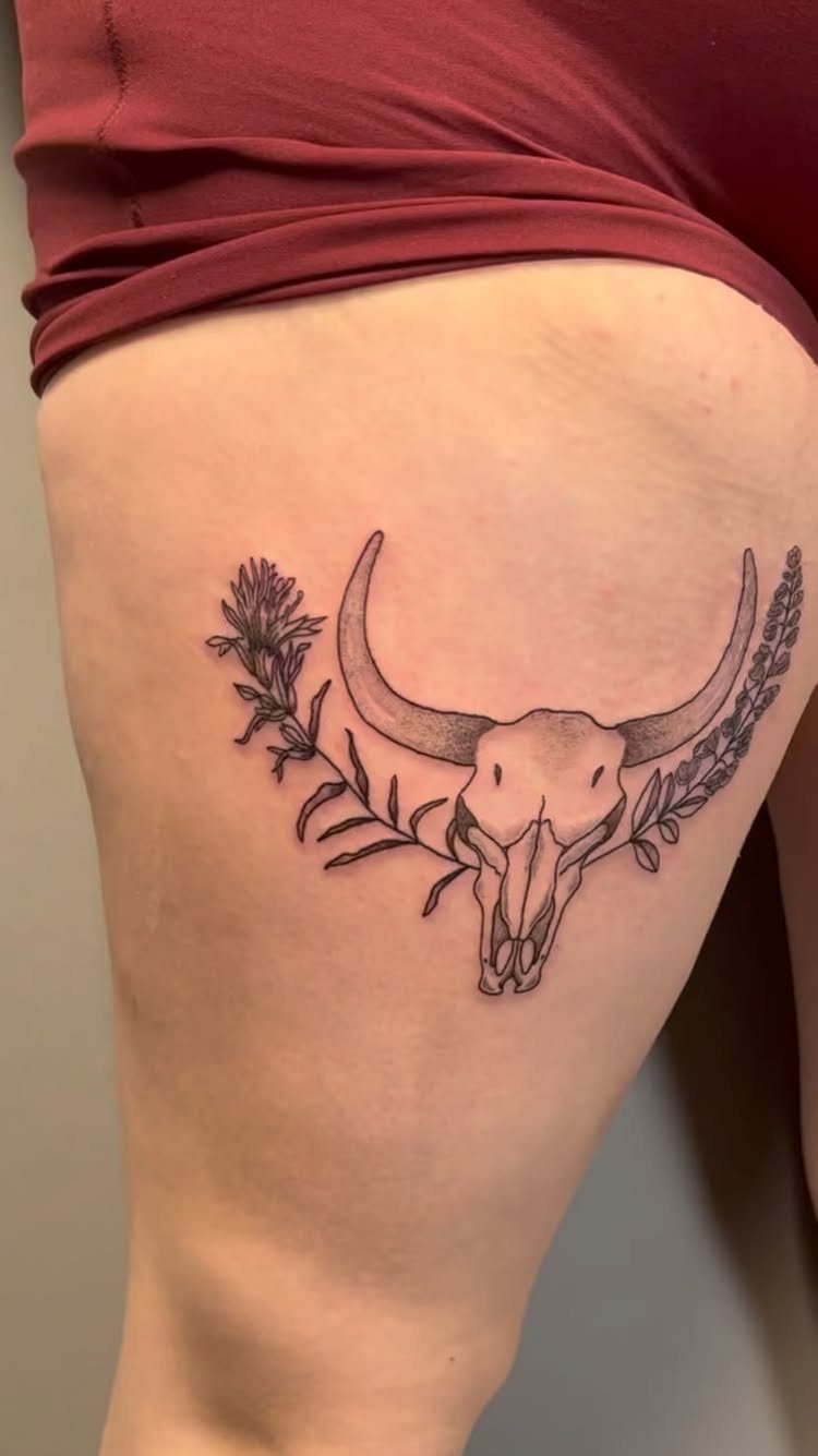 A black ink cow skull tattoo with leafy botanical branches on a person's thigh.