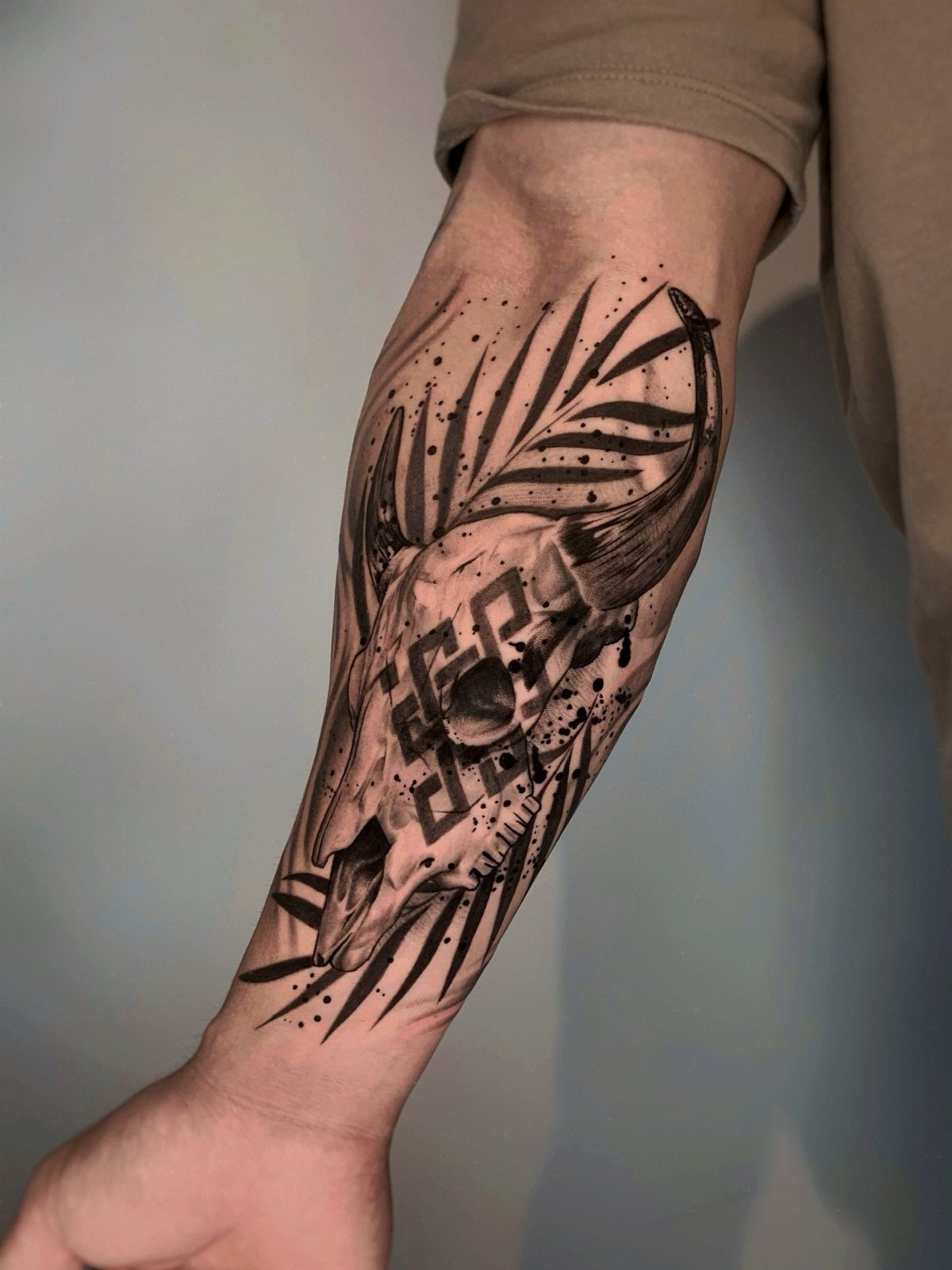 A black ink cow skull tattoo with geometric patterns and leaf shapes on a person's forearm.