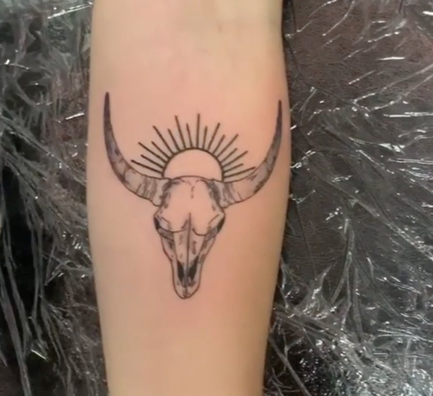 A black ink cow skull tattoo with a sunburst design on a person's arm.
