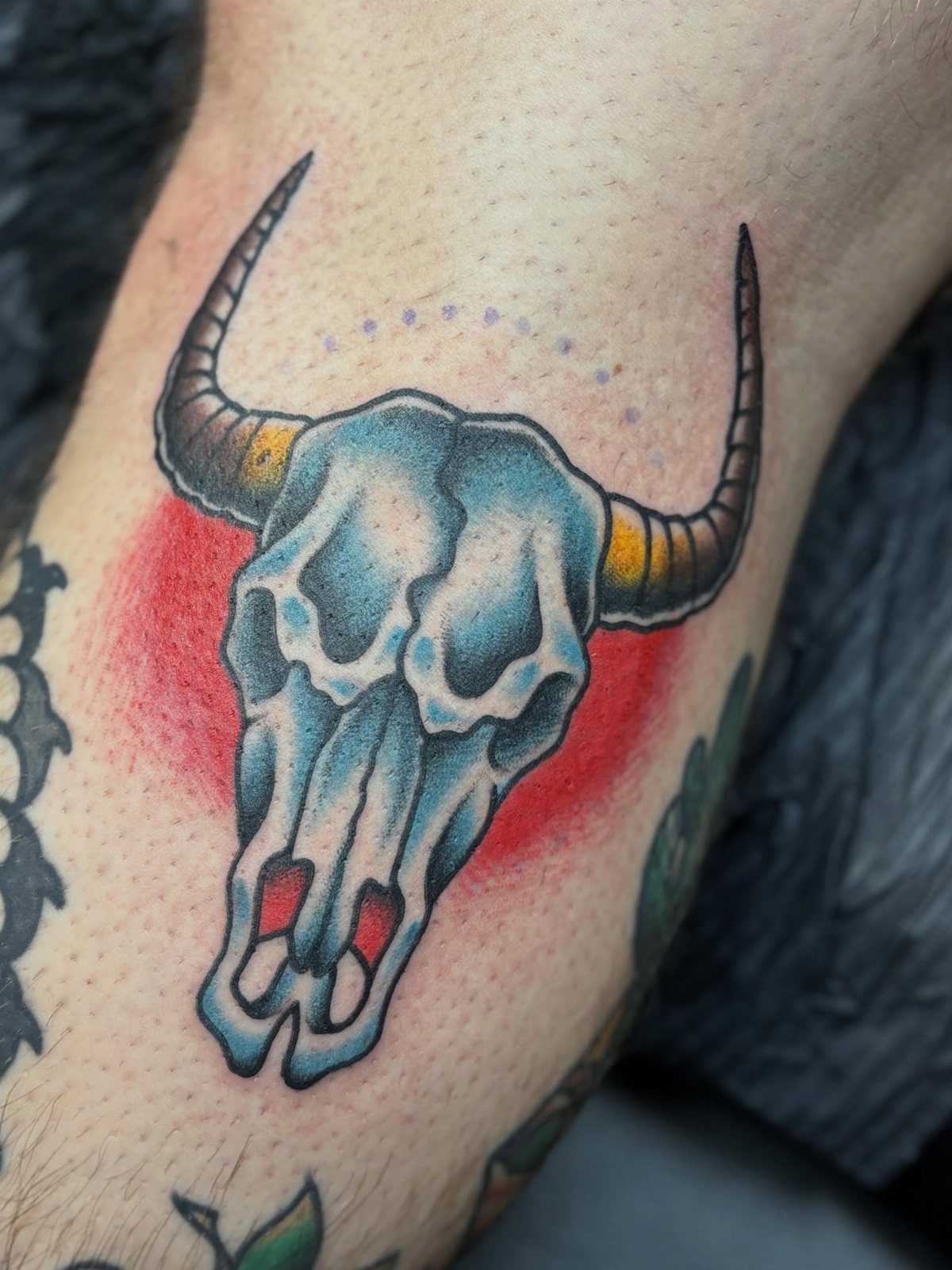 A colorful cow skull tattoo with blue shading and a red circular background on skin.