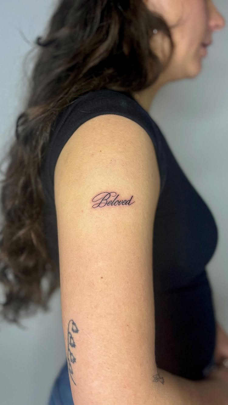 A woman's arm featuring a small cursive tattoo that reads the word Beloved.