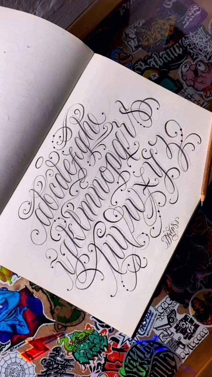 Hand-drawn decorative calligraphy alphabet with elaborate swirls in an open sketchbook on a desk.
