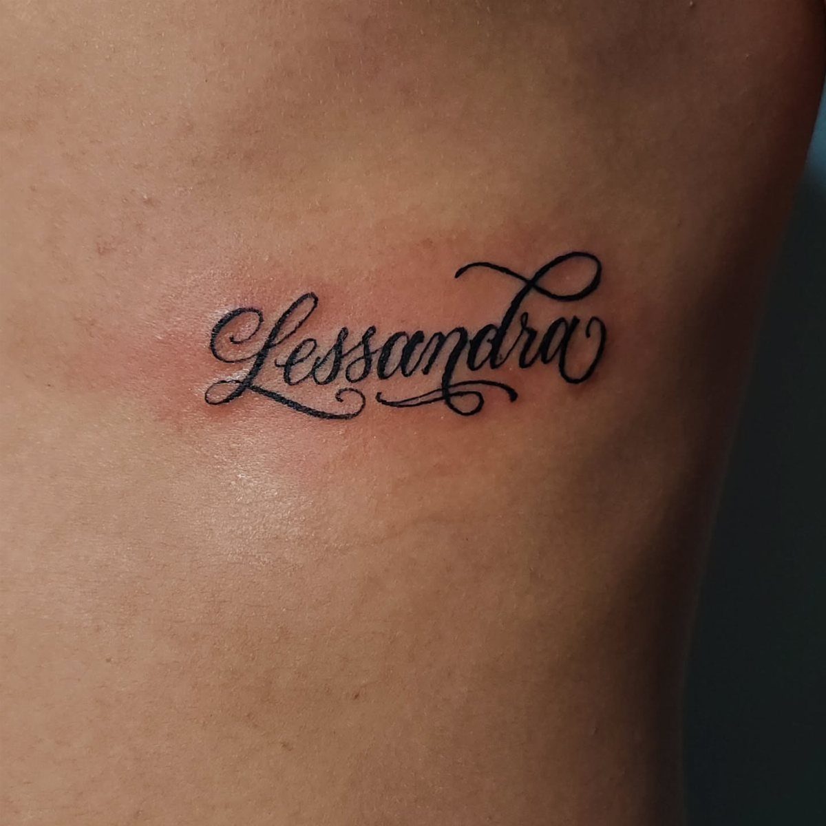 Cursive black ink tattoo of the name Lessandra on human skin.