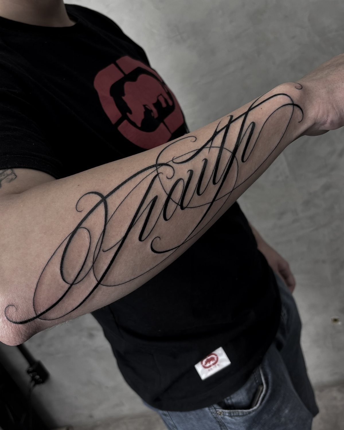 A large black ink cursive tattoo of the word Faith on a person's forearm.