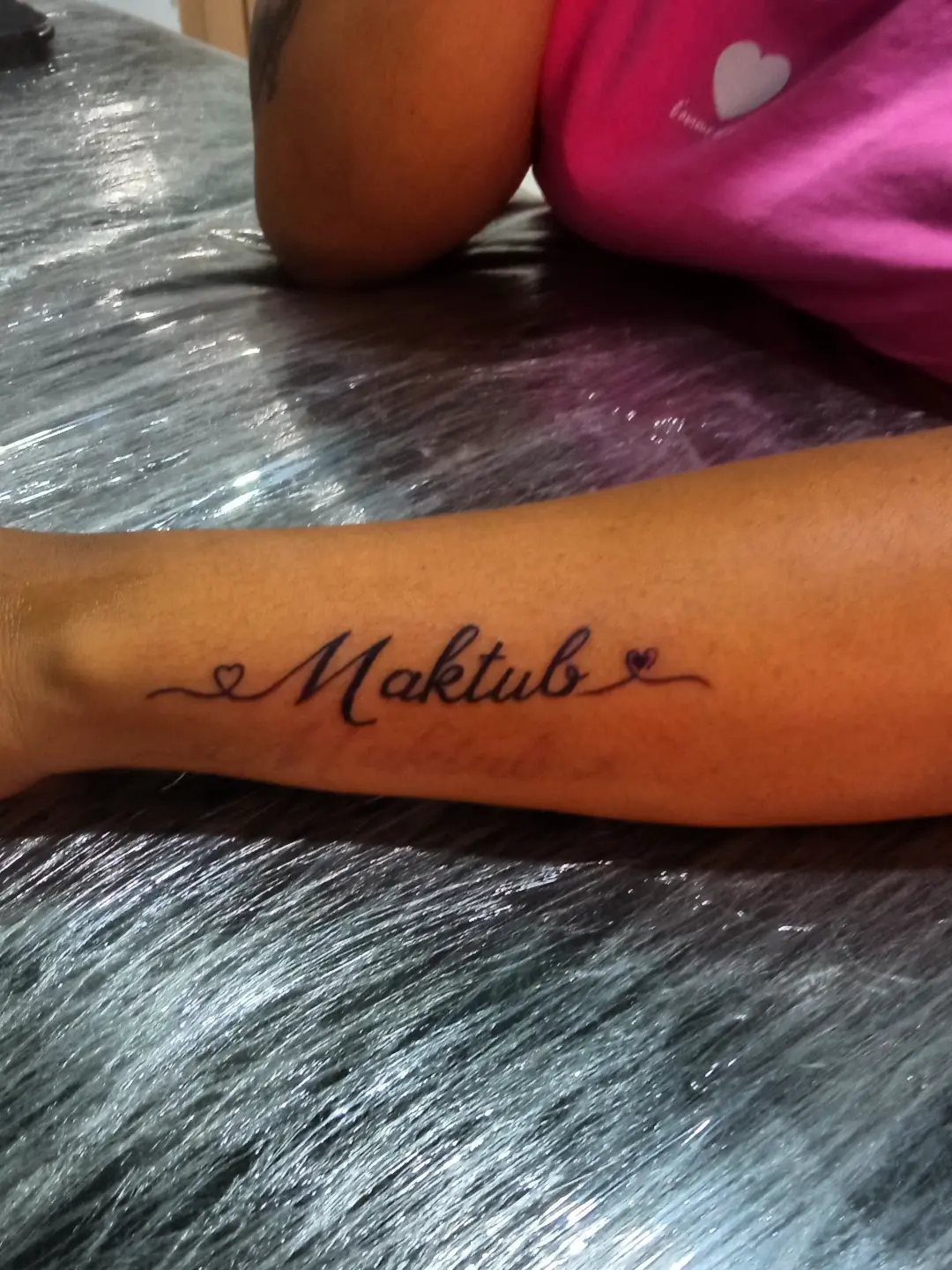 A forearm tattoo of the word Maktub in elegant black cursive script with small hearts.