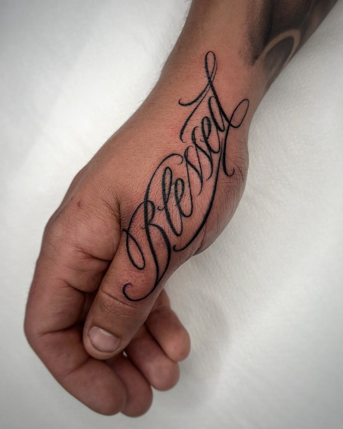 A hand with a black ink cursive tattoo of the word Blessed on the side.