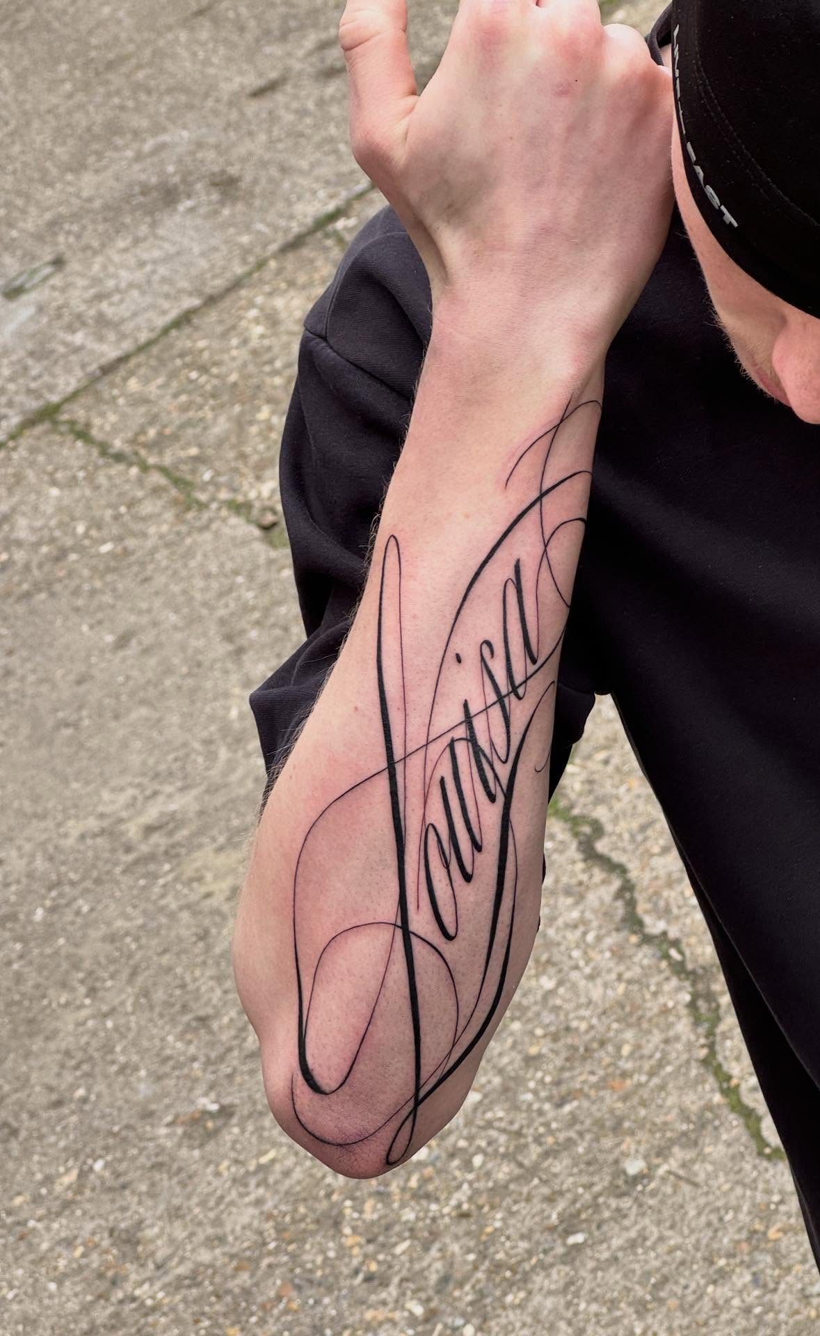 A forearm tattoo of the name Louisa written in elegant, flowing black cursive script.