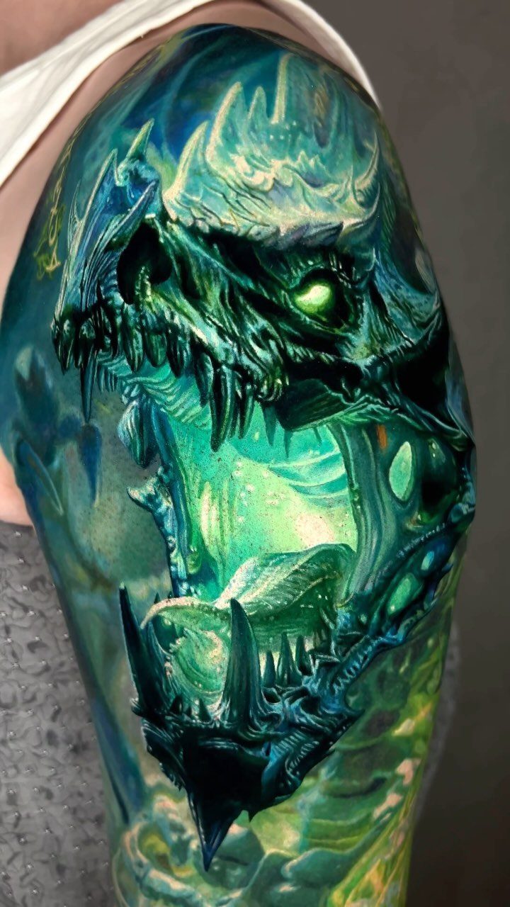 A detailed teal and green dragon skull tattoo with sharp teeth on a person's shoulder.