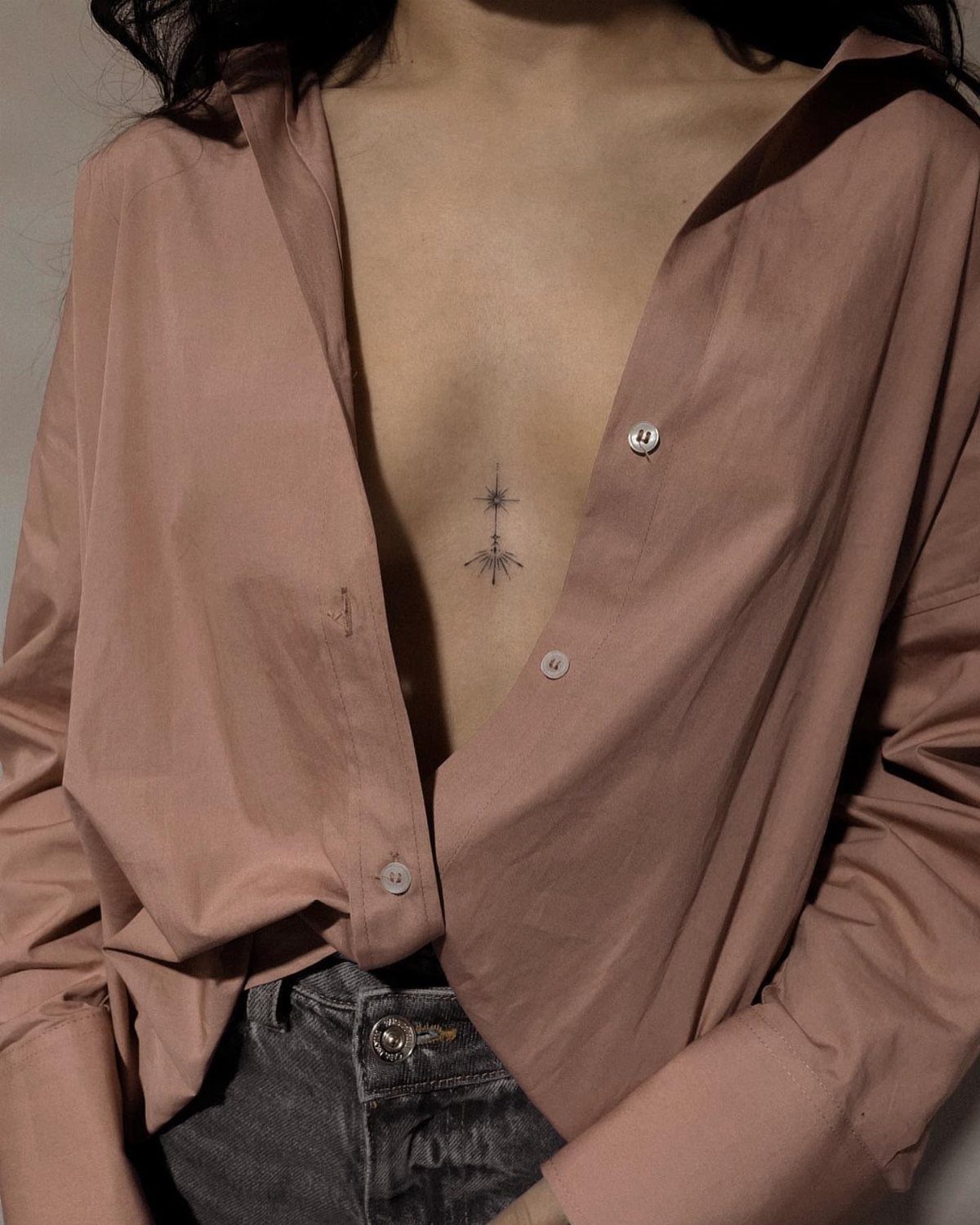 A small black starburst tattoo on a woman's sternum beneath an open pink shirt.