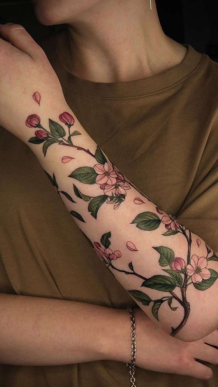 5 Fine Line Flower Vine Tattoo Ideas for Placement and Flow 2 A detailed botanical tattoo with pink blossoms and green leaves wrapping around a person's forearm.