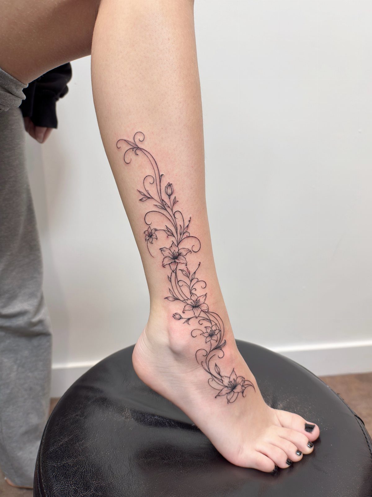 A fine line floral vine tattoo winding down a person's leg and foot.