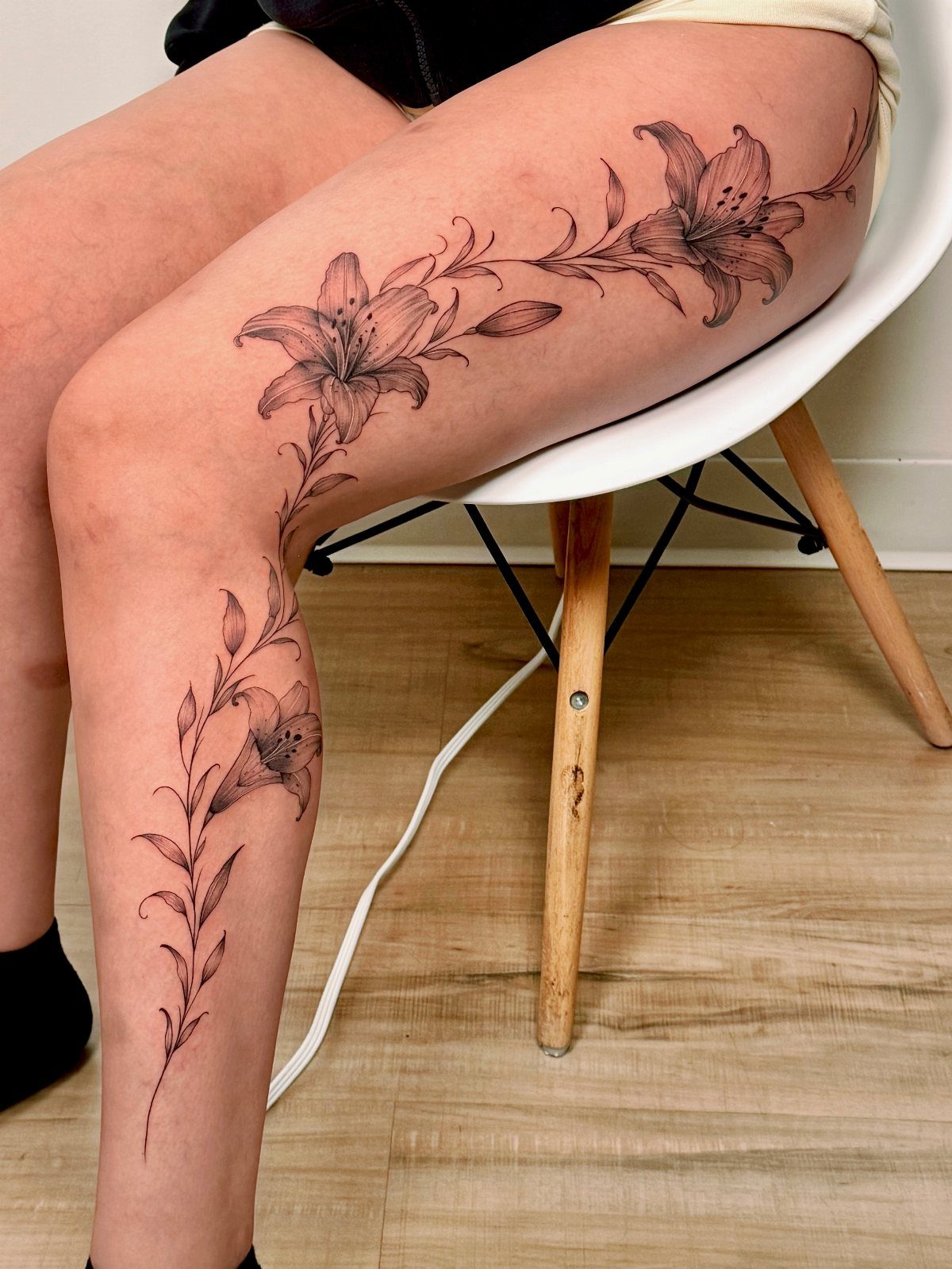 A black ink tattoo of large lilies and flowing vines wrapping around a person's leg.