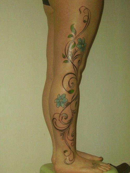 A leg with a large tattoo of swirling black vines, green leaves, and teal flowers.