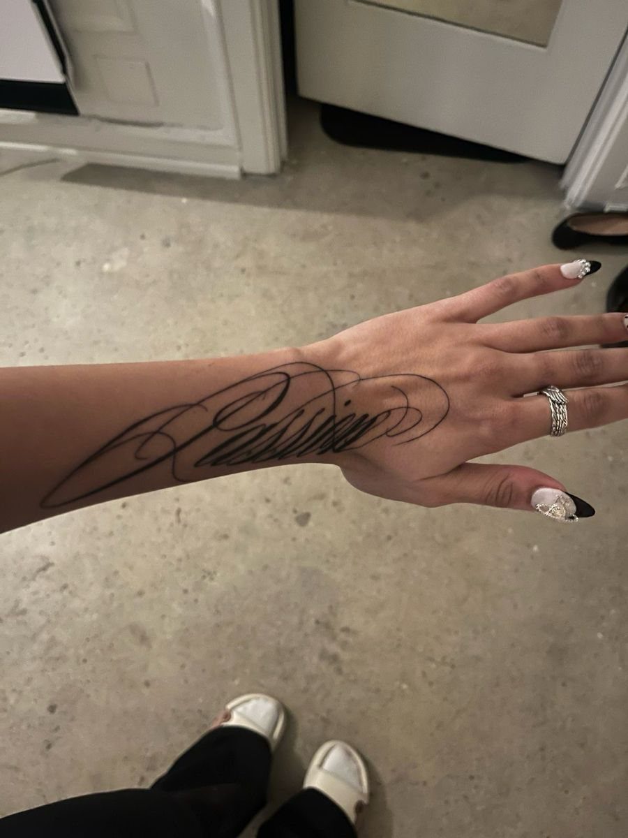 8 Forearm Cursive Tattoo Ideas for Script That Means Something 6 A person's forearm and hand featuring a large black cursive calligraphy design.