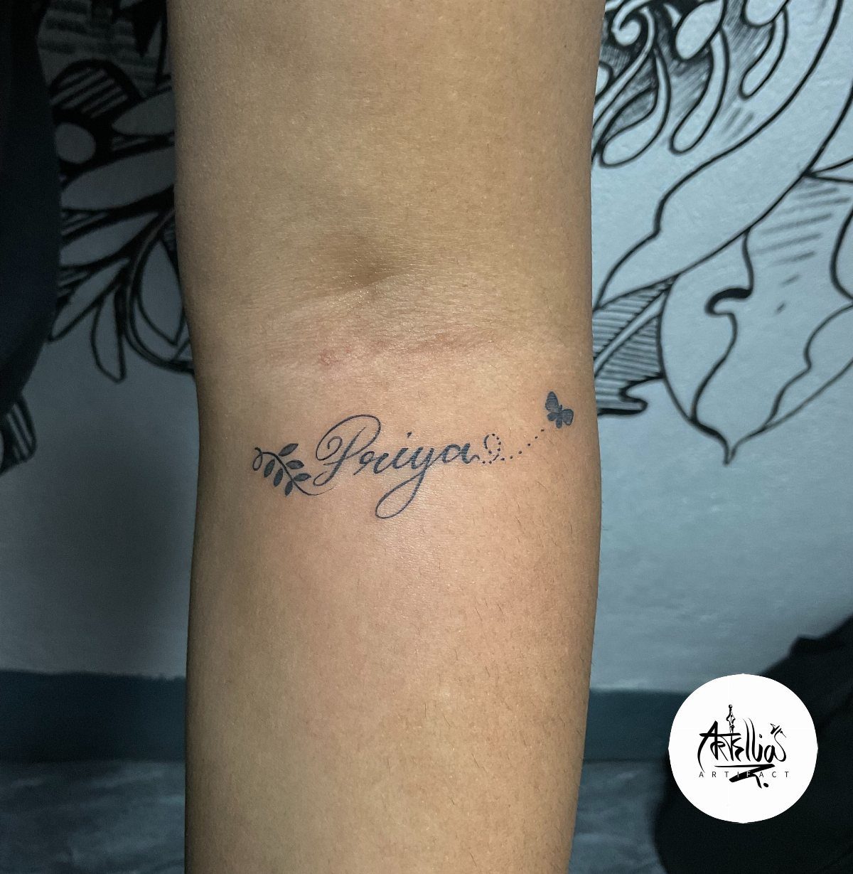 8 Forearm Cursive Tattoo Ideas for Script That Means Something 8 A cursive tattoo of the name Priya with a leaf and butterfly on a forearm.