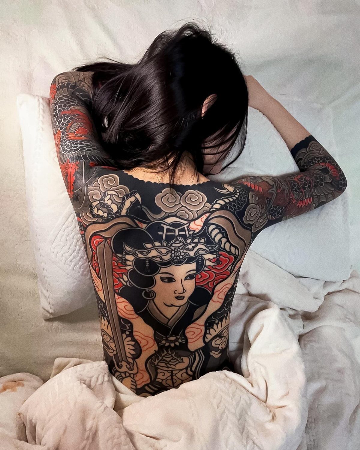 A person lying on a pillow displaying a large, colorful Japanese geisha backpiece tattoo.