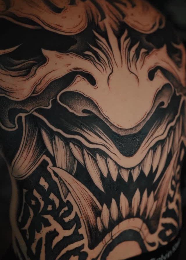 A detailed black and grey tattoo of a demonic face with sharp teeth and flowing lines.