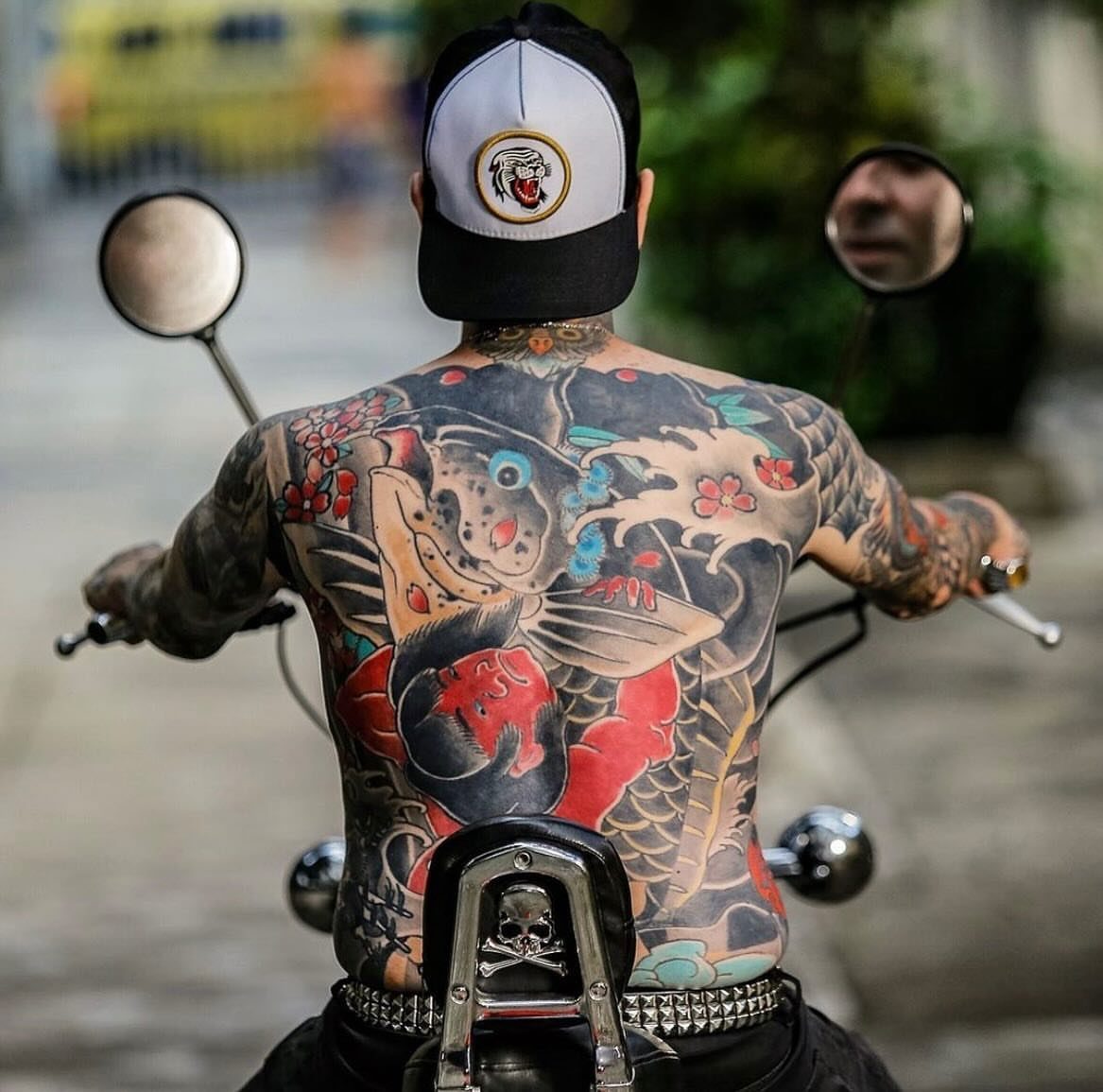 A man on a motorcycle displaying a large colorful koi fish backpiece tattoo.
