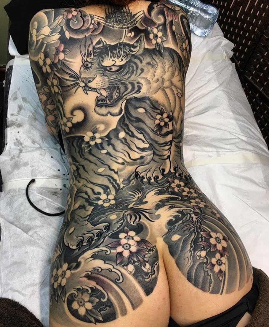 A large black and grey tiger tattoo with waves and flowers covering a person's back.