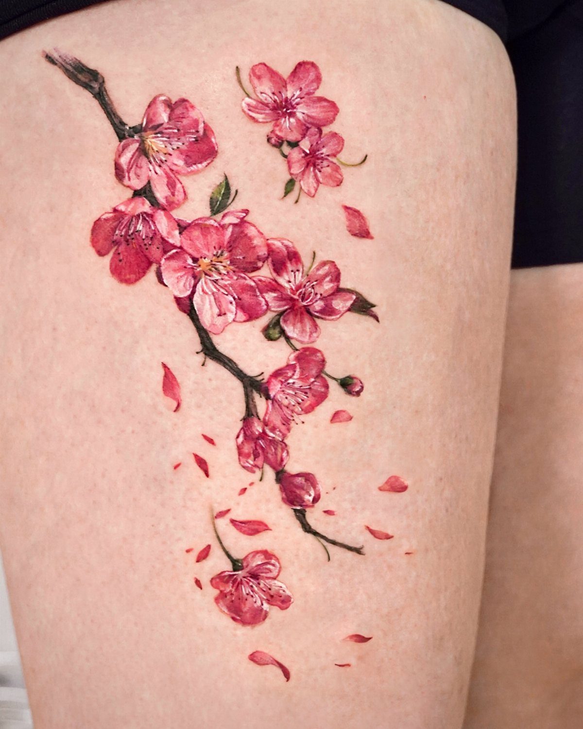 13 Large Flower Tattoo Ideas for Your Next Big Bloom 2 A large pink cherry blossom branch tattoo with falling petals on a person's thigh.