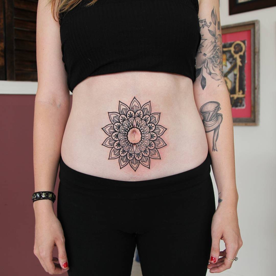 A woman's stomach featuring a black ink mandala tattoo centered on her navel.