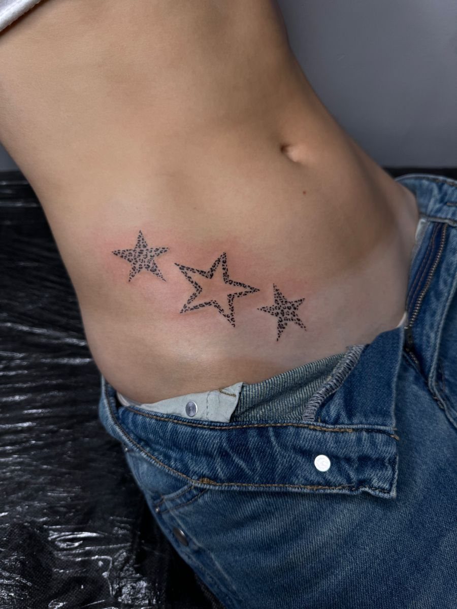 Three black dotwork star tattoos on a person's lower stomach area near blue jeans.