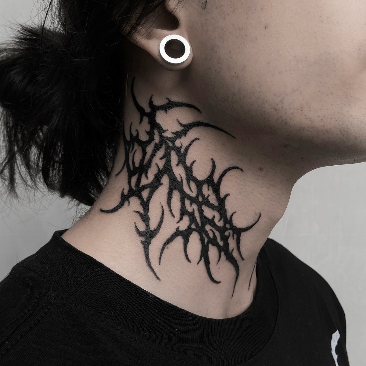 A person with a large black neo-tribal tattoo on their neck and a white ear gauge.
