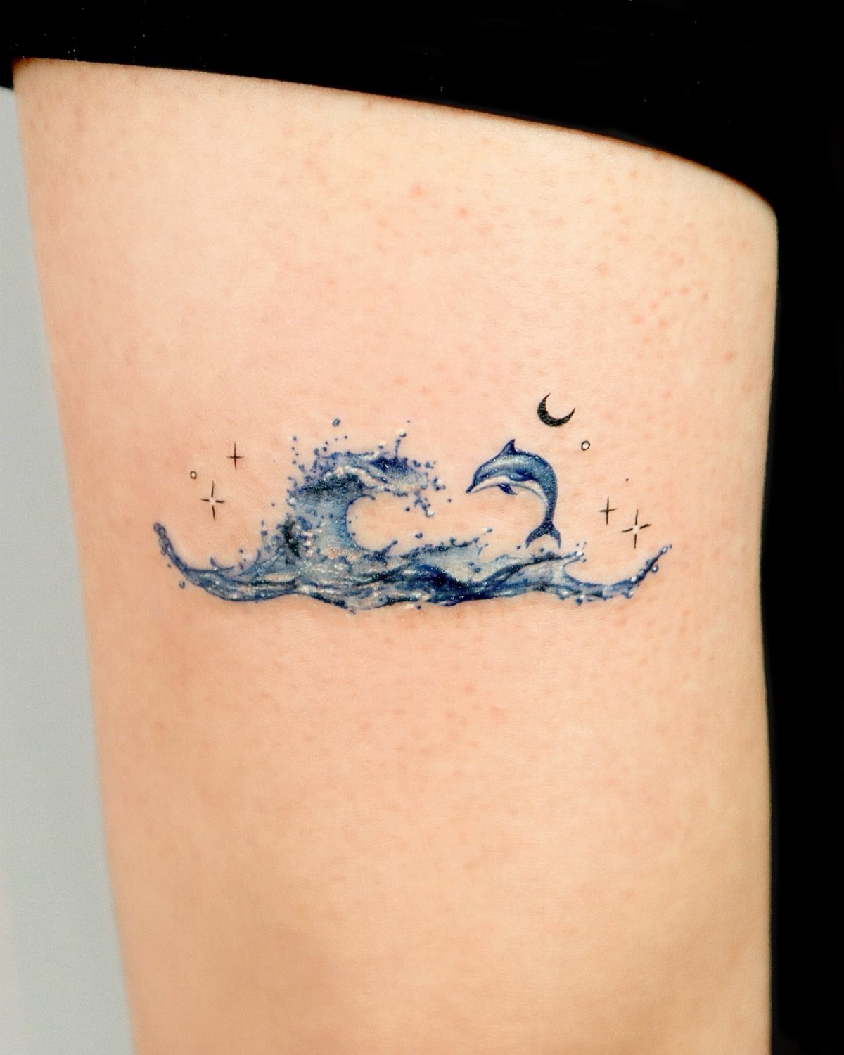 10 Ocean Themed Tattoos: From Fine Line Turtles to Black and Grey Mantas 8 A blue watercolor tattoo of a dolphin leaping through ocean waves on skin.