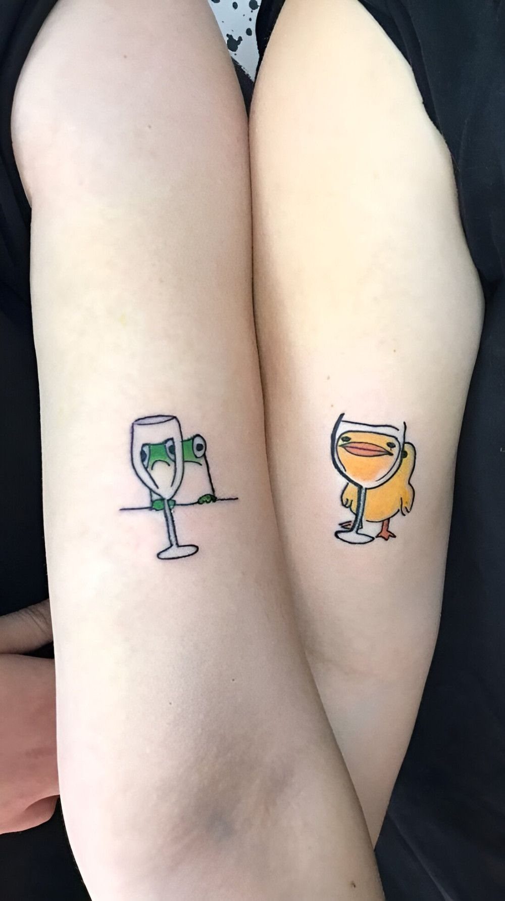Two small tattoos of a green frog and a yellow duck peeking out of wine glasses on forearms.