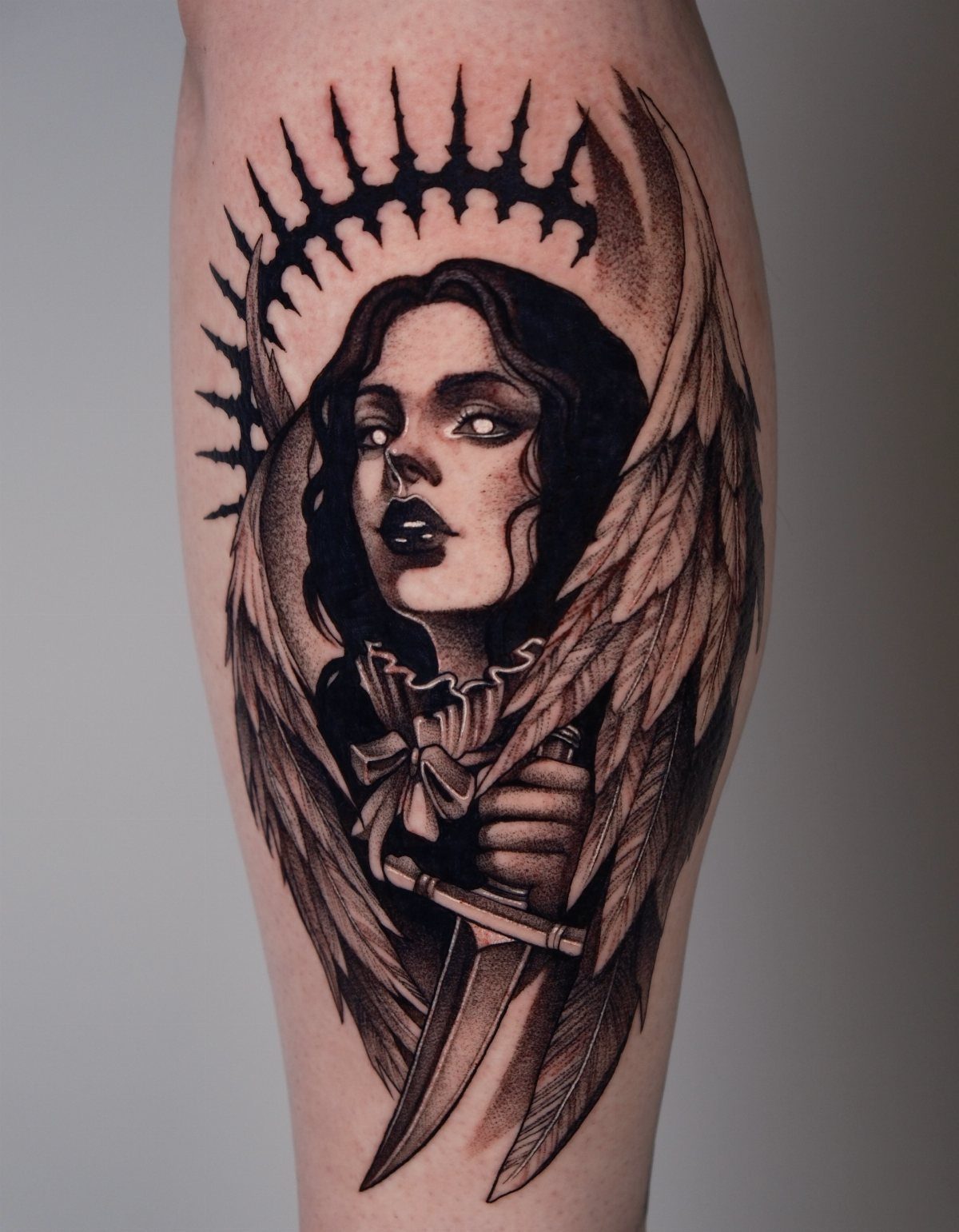 A black and grey gothic angel tattoo featuring a woman with wings and a spiked halo.