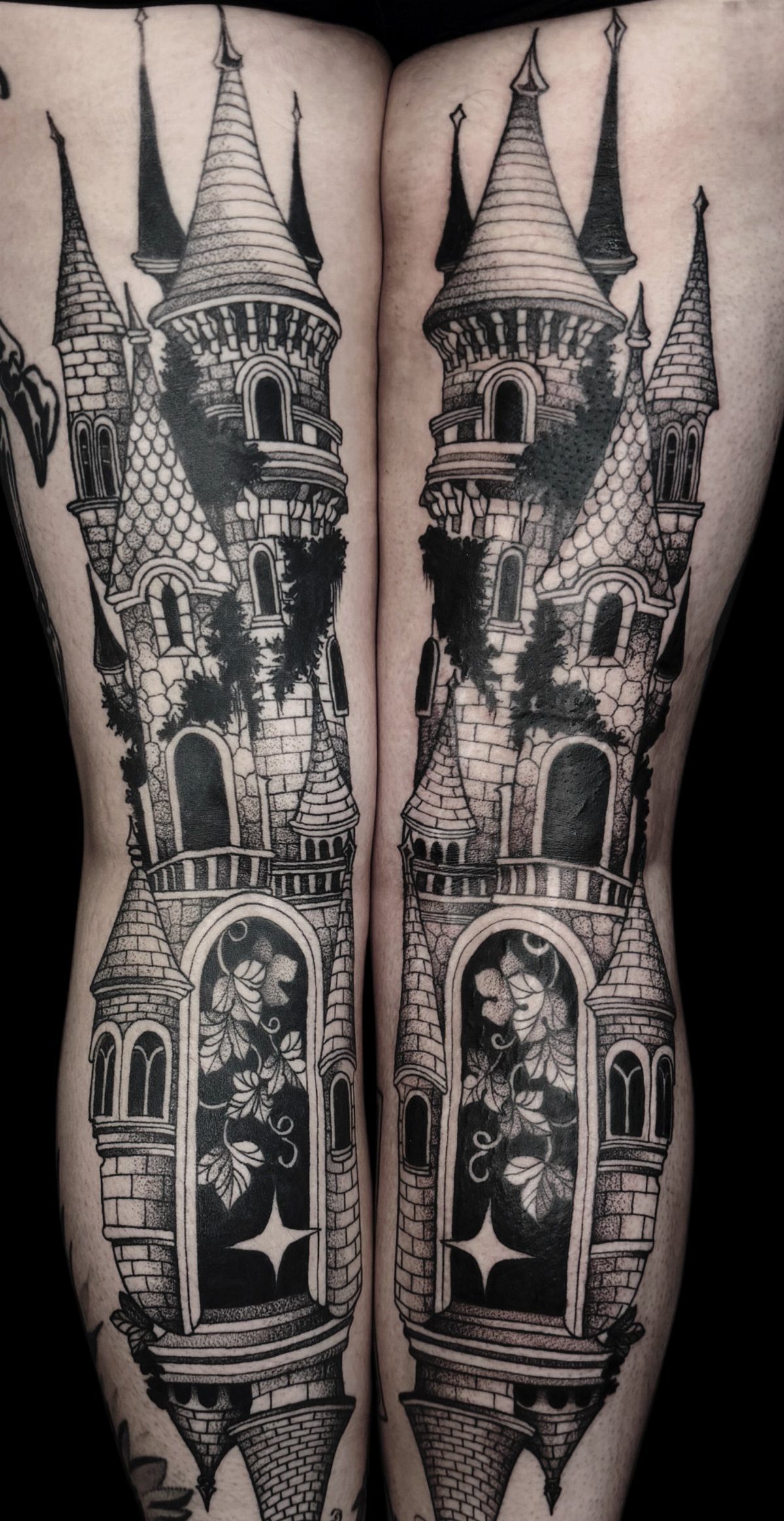 Two symmetrical gothic castle tattoos detailed in black and grey ink on a person's legs.
