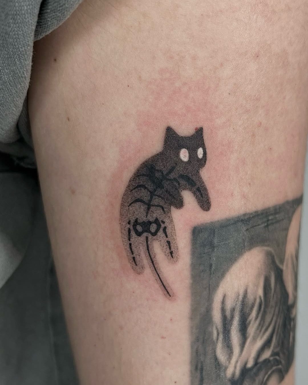 A small black ink tattoo of a spooky cat with white eyes on skin.