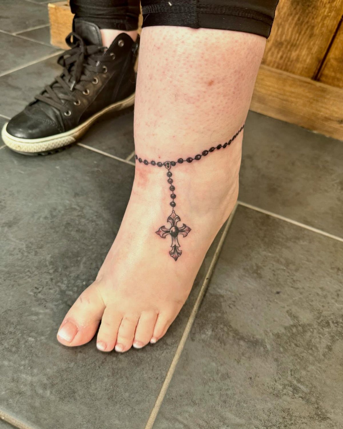 A black ink rosary tattoo with a cross centerpiece wrapped around a person's ankle.