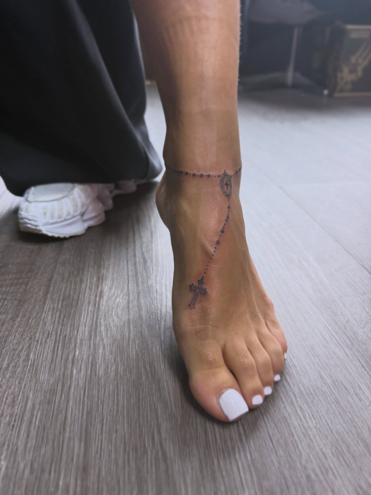 A fine-line rosary tattoo encircling a person's ankle and trailing down the foot.