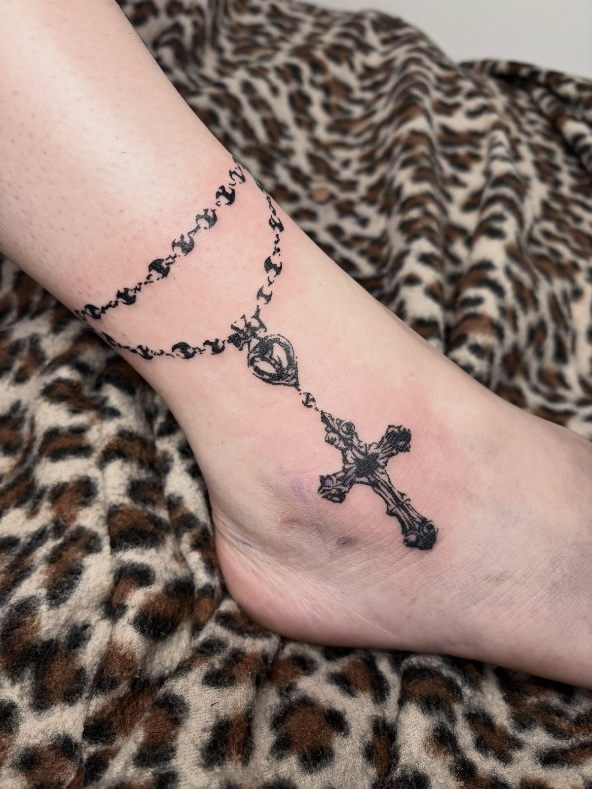 A black ink rosary tattoo with a heart centerpiece and gothic cross on a person's ankle.