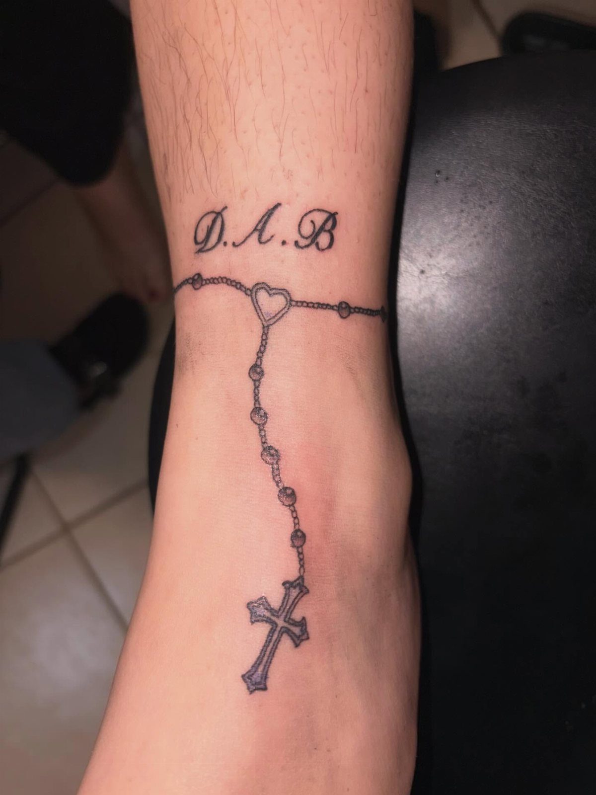 A rosary ankle tattoo featuring a heart centerpiece and a cross on a person's skin.