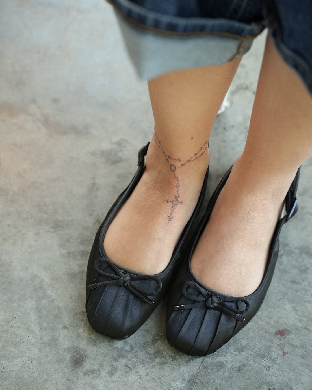 A person's ankles displaying a delicate fine-line rosary tattoo with a cross and black ballet flats.