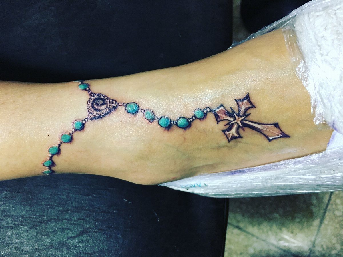 A turquoise bead rosary tattoo with an ornate cross wrapped around a person's ankle.
