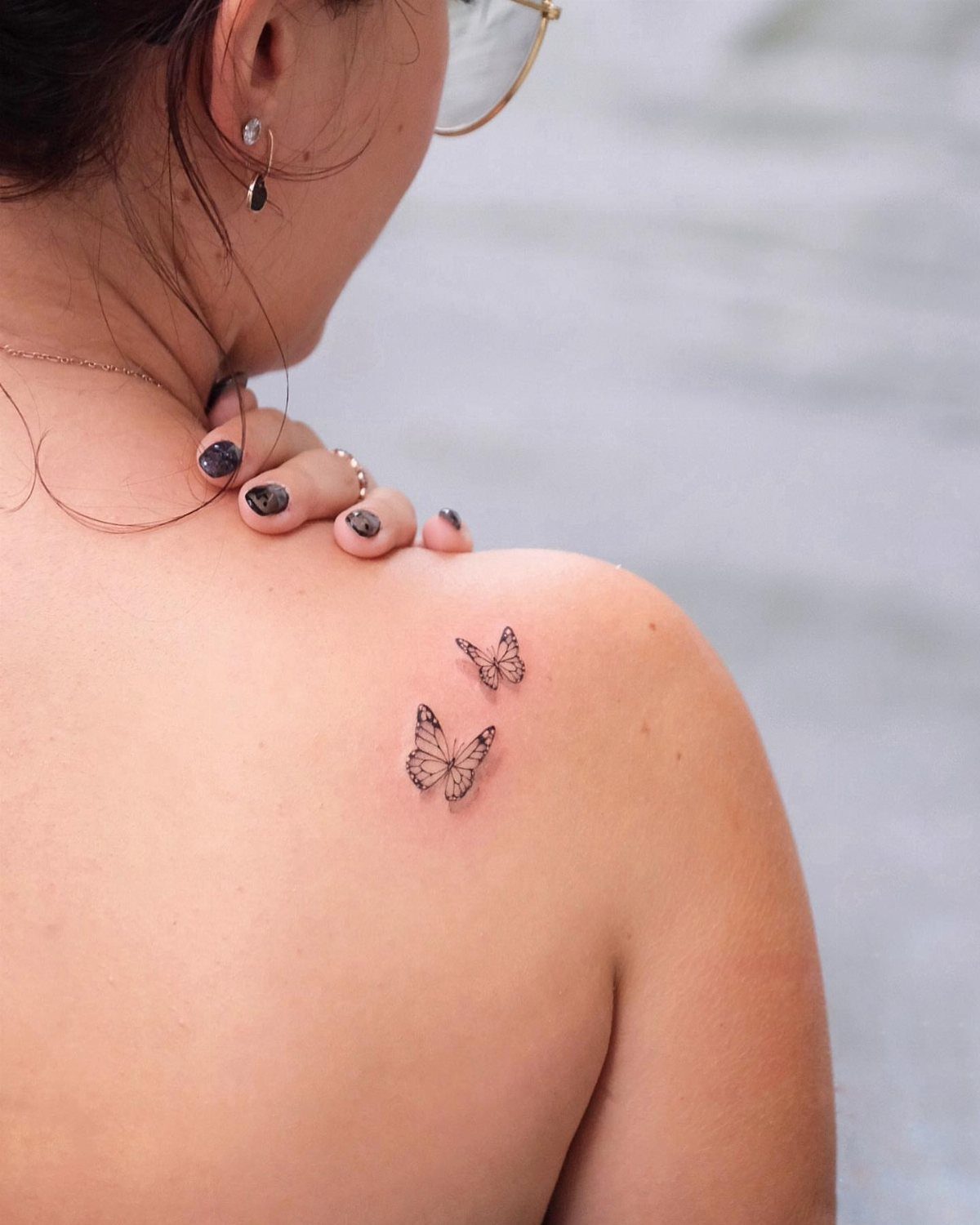 Two small fine line butterfly tattoos on a person's shoulder against a blurred background.