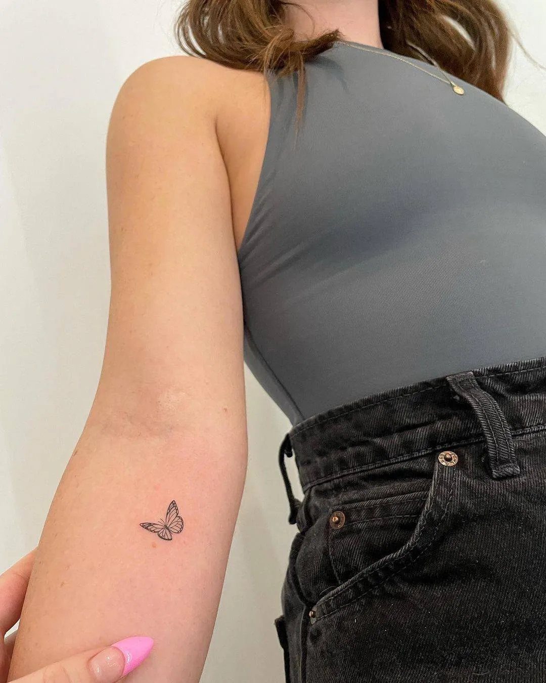 A tiny black butterfly tattoo on a person's inner forearm against a grey top.