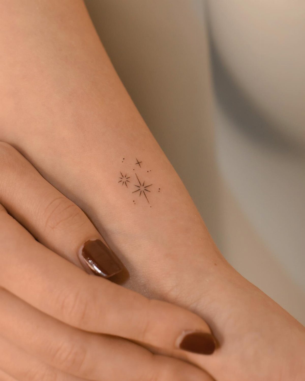 Small fine line star tattoos on a person's wrist with dark brown nail polish.