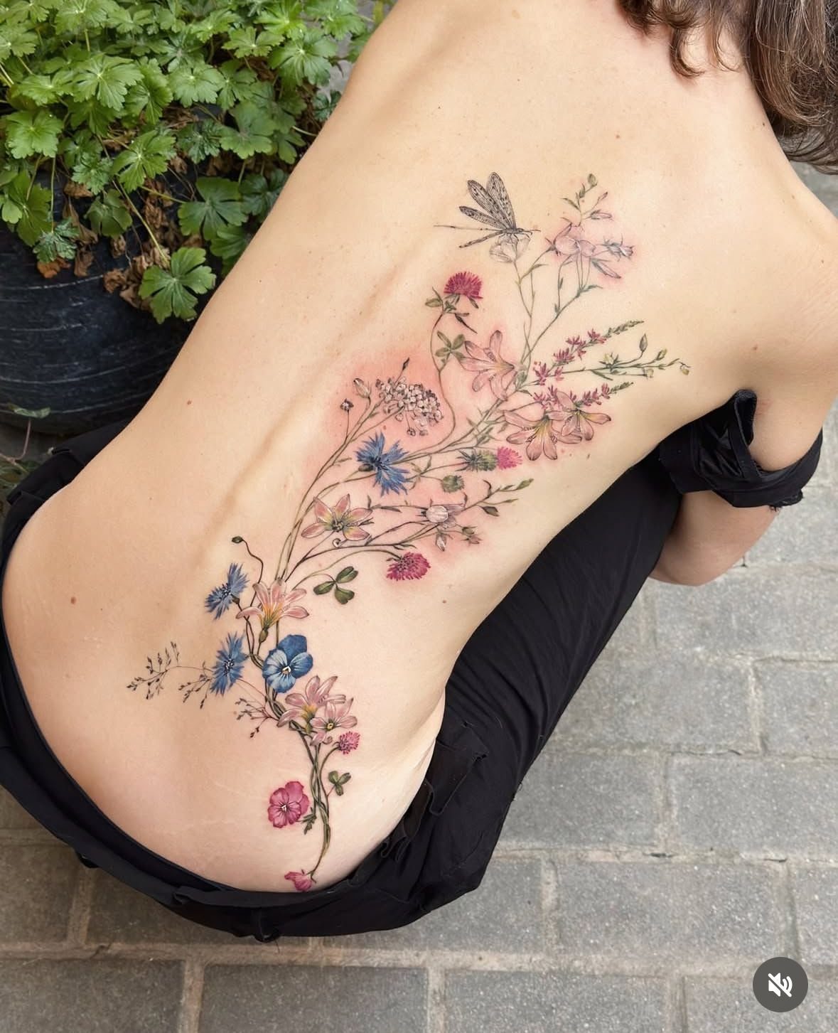 A woman's back featuring a colorful fine line wildflower and dragonfly tattoo along her spine.