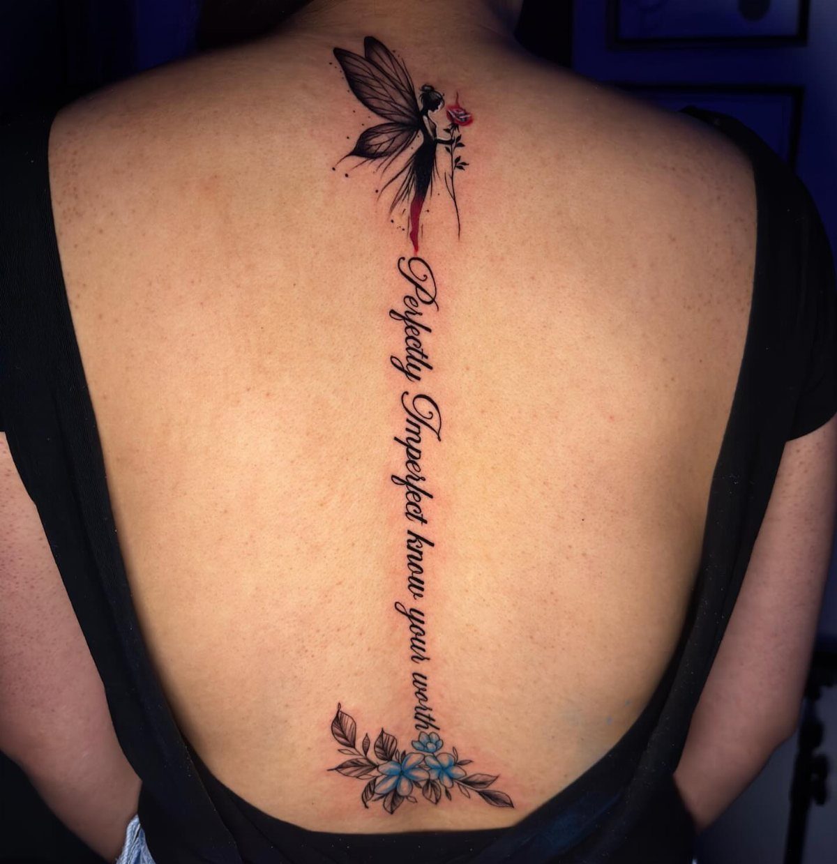 A woman's back featuring a vertical spine tattoo with a fairy, text, and blue flowers.